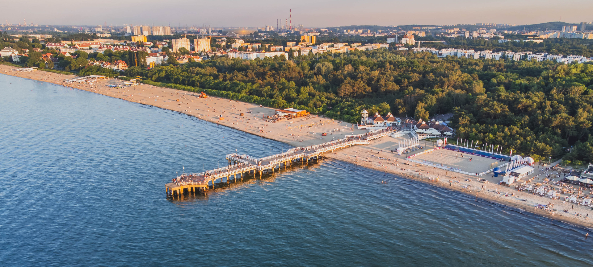 Widok z lotu ptaka na piaszczystą plażę z drewnianym molo, barami plażowymi, boiskami do siatkówki i miejską panoramą o zachodzie słońca
