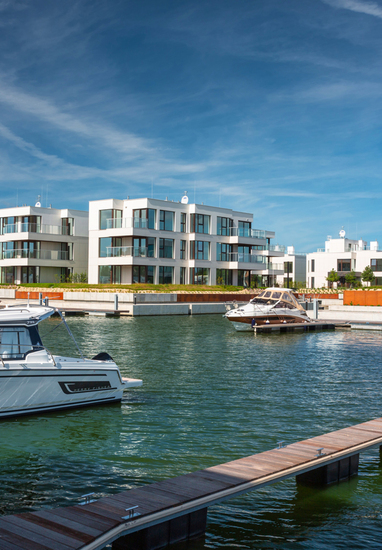 Modern white waterfront residences with glass balconies and private marina docks, yachts moored in a calm harbor.