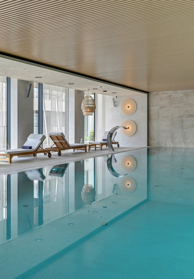 Relaxing indoor hotel pool with clear turquoise water, wooden loungers with white cushions, rattan pendants and a stainless water spout.