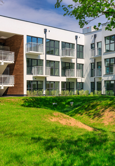 Modern three-story hotel with white façade, glass doors and private balconies, exterior stairwell and green lawn.