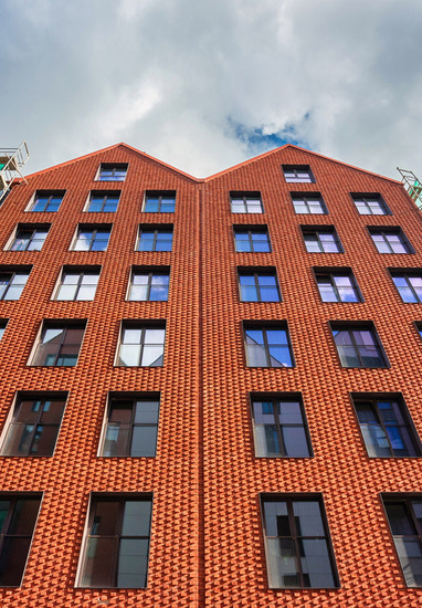 Modern red-brick hotel façade with repeating windows and gabled roof, flanked by green scaffolding netting during renovation under a cloudy sky