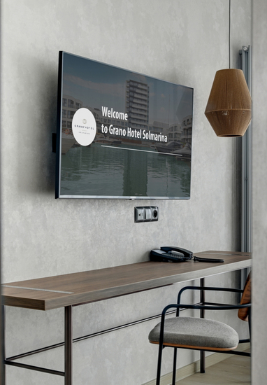 Modern hotel workspace with wall TV showing welcome screen, wooden desk with phone, chair, pendant lamp, bed and curtains.
