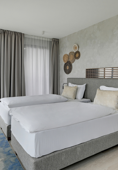 Modern twin hotel room with upholstered headboard, woven pendant lights, wooden side table, desk and large window view.