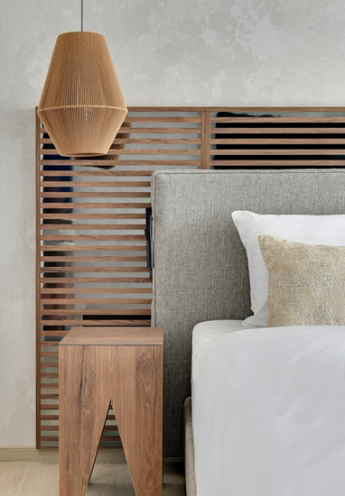 Hotel bedroom corner: gray upholstered headboard, white linens, beige pillow, slatted wood screen, bedside table and woven lamp.