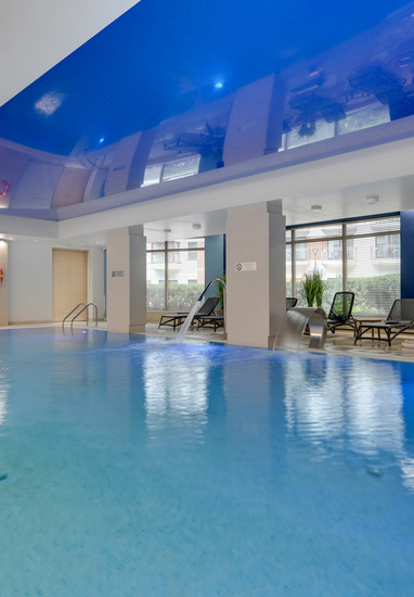 Indoor hotel pool with glossy blue reflective ceiling, lounge chairs, cascading water jets, stainless handrails and large windows.