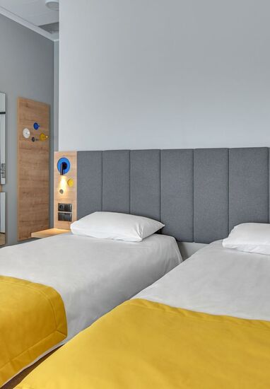 Twin beds with grey upholstered headboard, bright yellow bed runners, bedside reading lights, built-in power outlets, wardrobe and full-length mirror