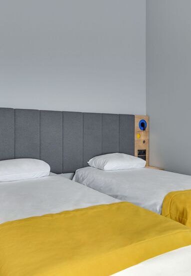 Twin beds with yellow throws, upholstered grey headboard and wooden bedside panels with reading lights in a clean modern hotel room.