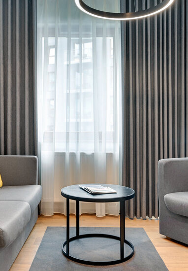 Hotel lounge with grey sofa and armchair, yellow cushions, round black coffee table and sheer plus dark drapes.