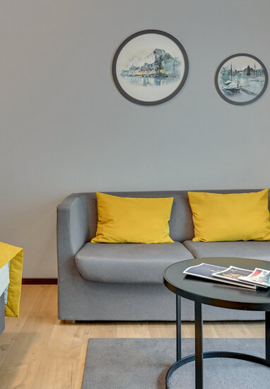 Modern guestroom with grey sofa and two yellow cushions, matching yellow bed runner, round black coffee table with magazines and circular wall art