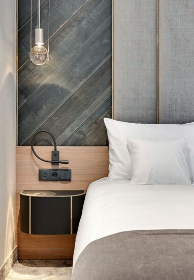 Modern hotel bedside with white linens, padded grey headboard, diagonal dark-wood accent panel, glass pendant, flexible reading light and wall shelf.