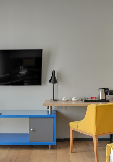 Wall-mounted flat-screen TV above a work desk with electric kettle, cups on a tray, black floor lamp and yellow chair
