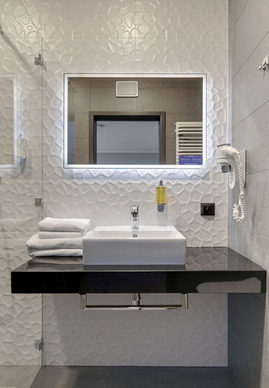 Modern hotel bathroom with glass shower, illuminated mirror, white basin on black counter, stacked towels and hairdryer.