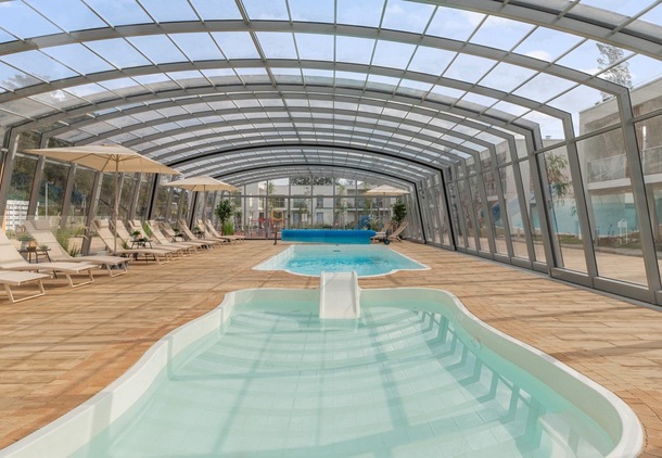 Covered indoor pool complex with glass arched roof, two pools including a shallow kids' pool with slide, lounge chairs and umbrellas.