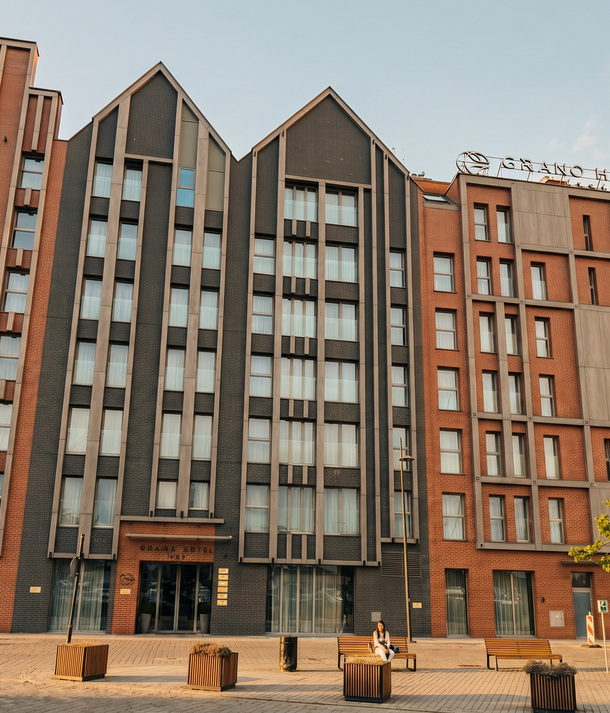 Modern brick hotel with peaked gables and tall windows, ground-floor entrance, benches and a guest sitting outside under warm sunset light.