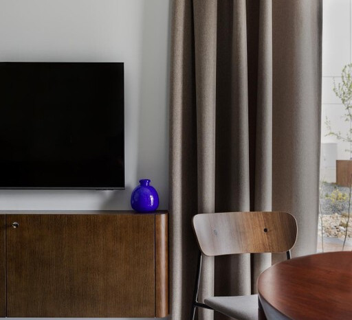 Hotelowy kącik wypoczynkowy z telewizorem na ścianie, drewnianą szafką i krzesłem, okrągłym stołem, zasłonami i niebieską wazą.