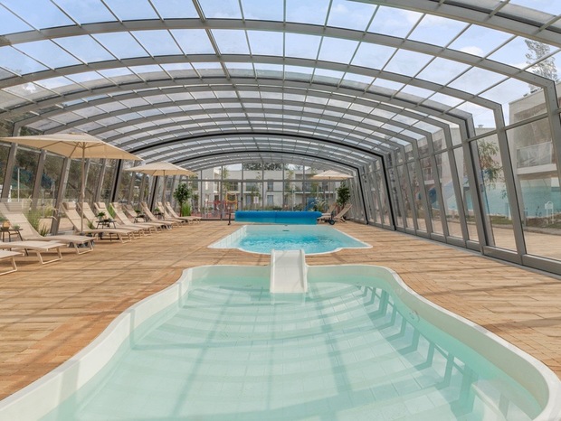 Covered indoor pool complex with glass arched roof, two pools including a shallow kids' pool with slide, lounge chairs and umbrellas.