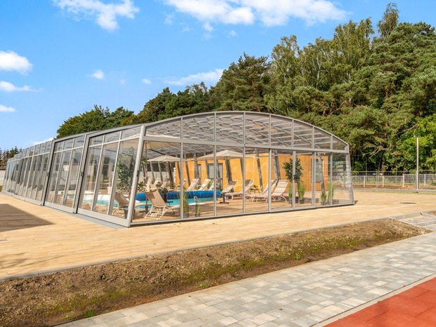 Glass-enclosed pool pavilion with loungers on a wooden deck, indoor heated pool and plants, set beside a forest under a blue sky