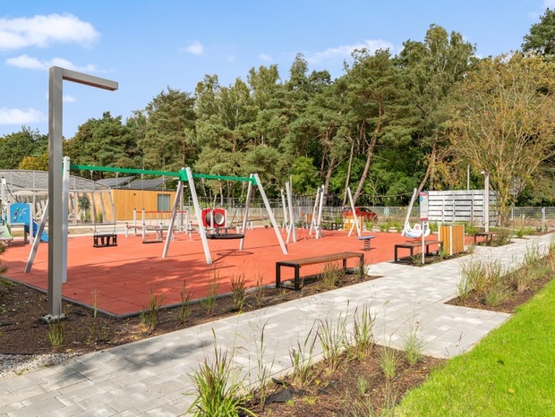 Family-friendly outdoor playground with swings and benches on red rubber safety surface, paved path and forested backdrop