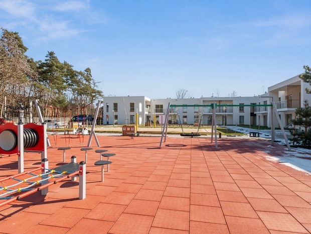 Outdoor family playground with swings and climbing equipment on red safety tiles, set before modern white hotel rooms