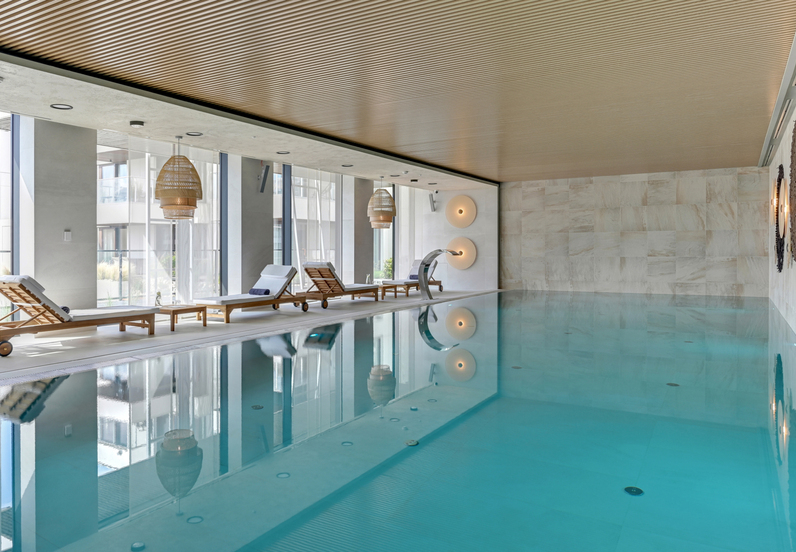 Relaxing indoor hotel pool with clear turquoise water, wooden loungers with white cushions, rattan pendants and a stainless water spout.