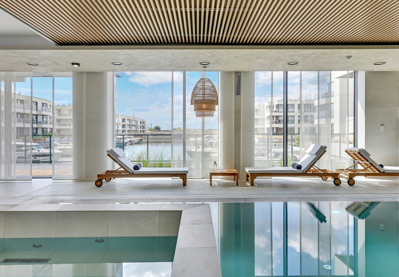 Indoor pool lounge with teak sunbeds and rolled towels, floor-to-ceiling windows overlooking marina and modern wood-slat ceiling.