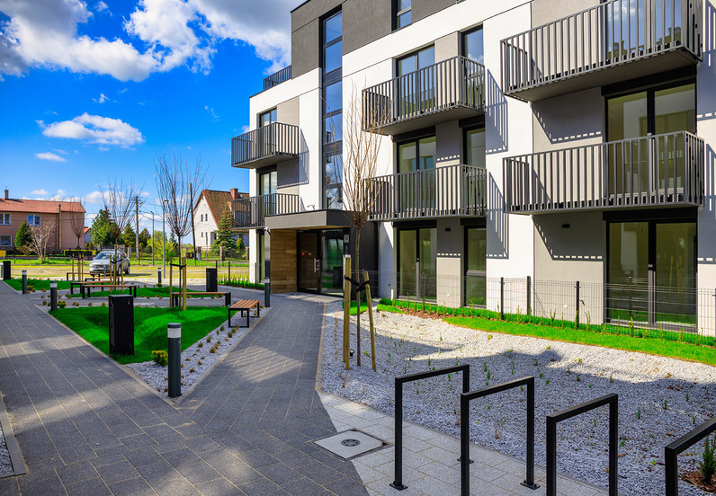 Modern hotel façade with private balconies facing a landscaped courtyard, paved walkways, benches, bike racks and small garden beds under a bright blue sky