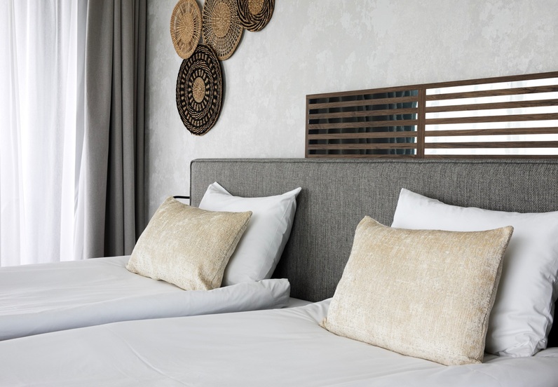 Twin beds with white linens and beige cushions, grey upholstered headboard with slatted wood panel, woven wall decor and sheer curtains