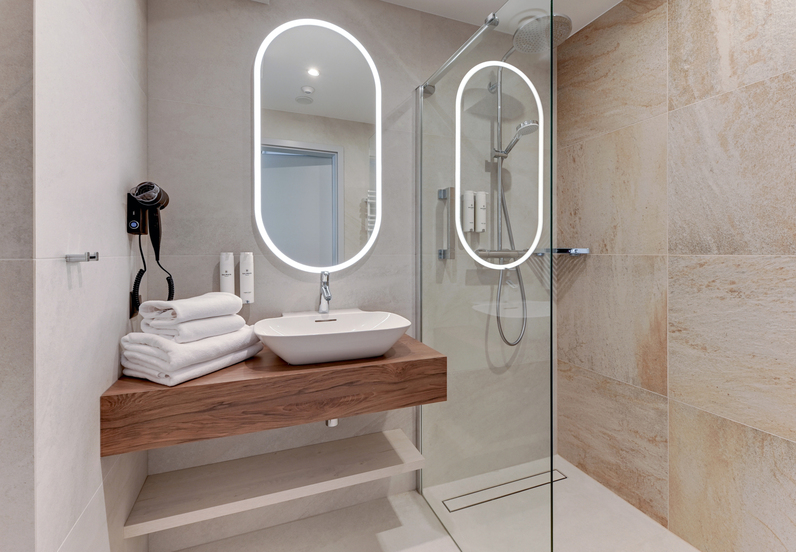 Modern hotel bathroom with illuminated oval mirror, vessel sink on wooden floating vanity, stacked towels, hairdryer and glass walk-in shower.
