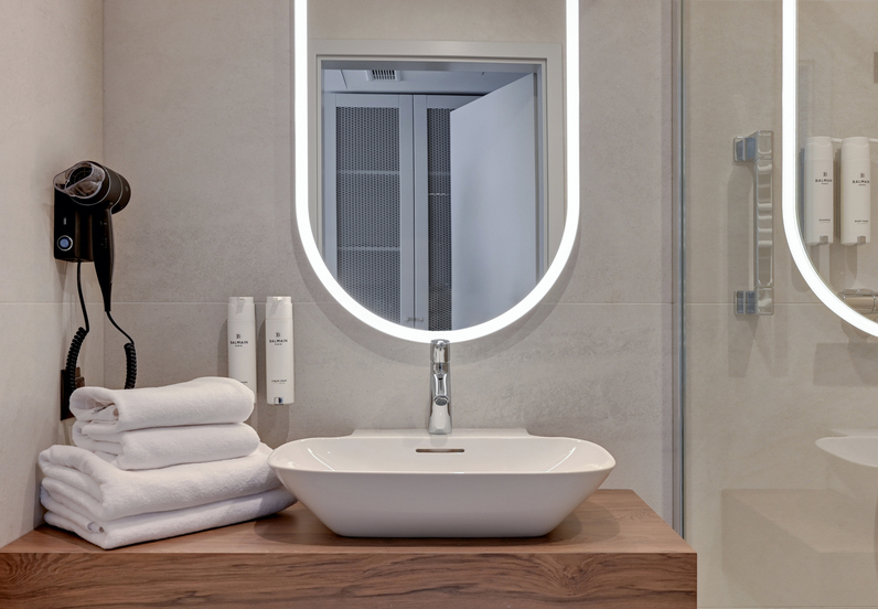 Hotel bathroom with LED oval mirror, vessel sink on wooden vanity, stacked white towels, wall hairdryer and soap dispensers.