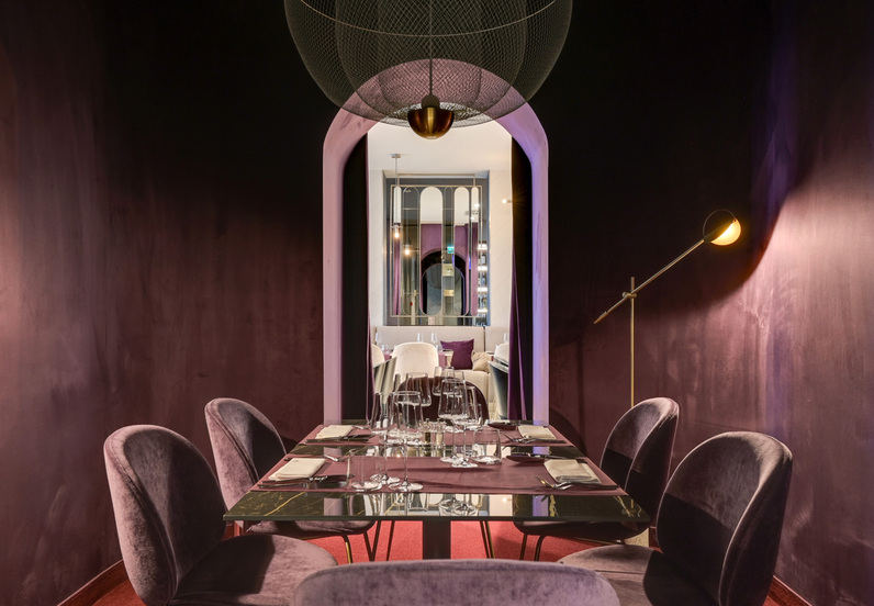 Przytulna prywatna sala restauracyjna z fioletowymi welurowymi krzesłami, marmurowym stołem nakrytym na kolację i łukowym przejściem.