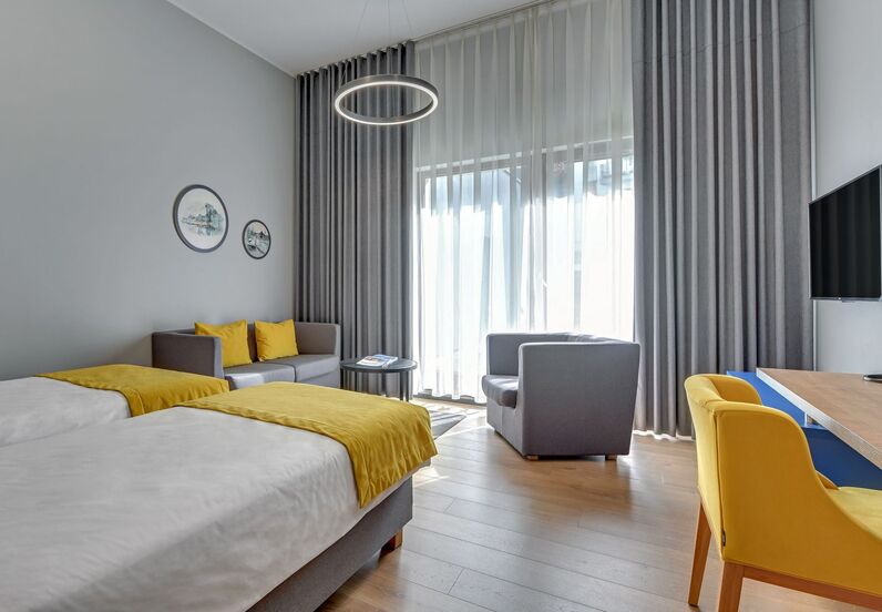 Modern twin hotel room with yellow accents, sofa and armchair seating, desk and wall TV, large sheer curtains letting in soft daylight.