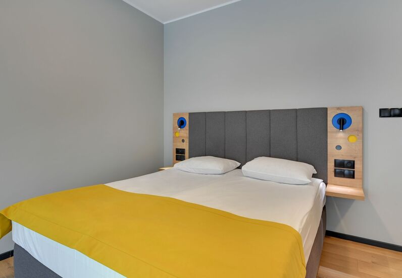 Modern double bed with grey upholstered headboard, crisp white linens, yellow bed runner, bedside reading lights and power outlets.