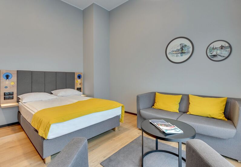 Contemporary hotel room with double bed, grey headboard, yellow bed runner, sofa with yellow cushions and coffee table.