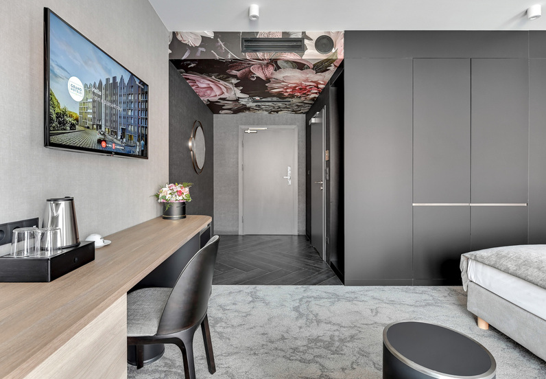 Modern hotel room with desk, kettle and glasses, wall TV, floral-patterned ceiling above entry, large wardrobe and bed.