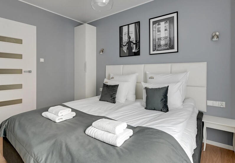 Modern double hotel room with king bed, white headboard, grey linens, folded towels on bed, bedside table, wardrobe and framed wall art.