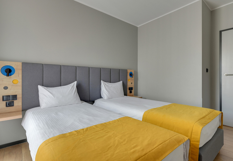 Two twin beds with grey upholstered headboard, yellow bed runners, bedside reading lamps and built-in power outlets.