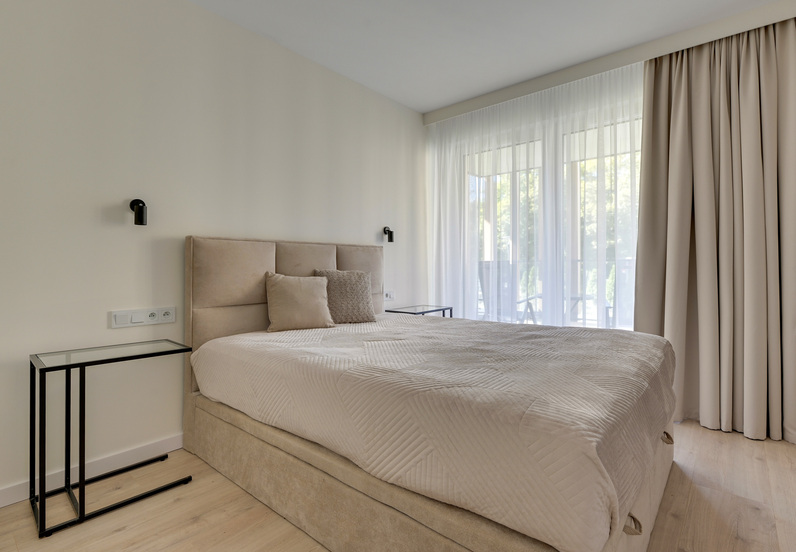 Jasny nowoczesny pokój hotelowy z beżowym tapicerowanym łóżkiem, pościelą, lampkami przy łóżku i balkonem z zasłonami.
