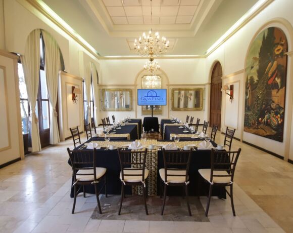 Sala de conferencias elegante con techos altos, candelabros, largas mesas con cristalería y servilletas blancas, y arte decorativo en la pared