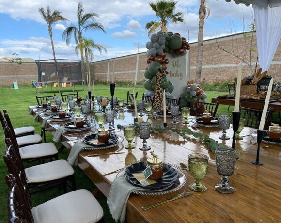 Elegante montaje al aire libre para baby shower con mesa larga de madera decorada, globos verdes y marrones y palmeras bajo cielo azul