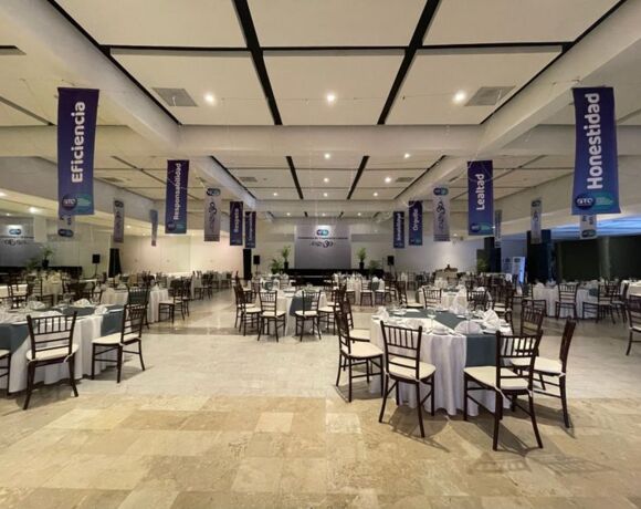 Amplio salón de banquetes con mesas redondas preparadas para evento formal, manteles blancos, sillas de madera y pancartas en el techo