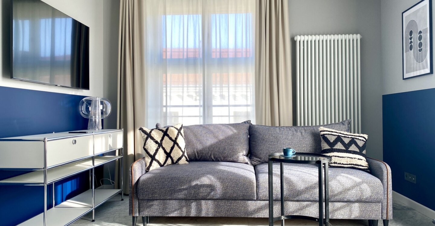 Jasny salon w apartamencie. Pośrodku szara kanapa z poduszkami. Przed nią stolik kawowy. Na ścianie po lewej telewizor, a pod nim biała szafka. z tyłu okno z firankami i zasłonami.