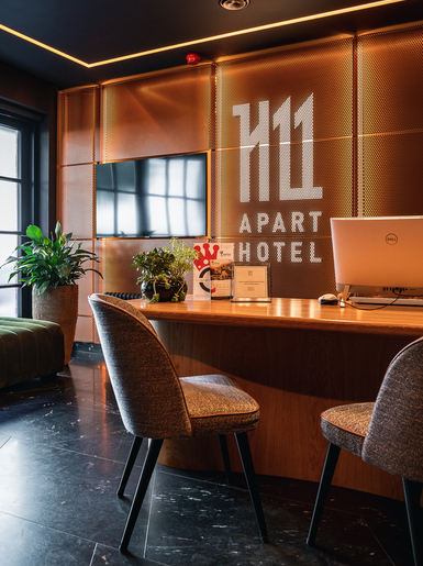 On the reception desk sits a bright monitor and a plant in a black pot. In front of it are two gray upholstered chairs. Further back, to the left, is a green sofa, and next to it is a plant in a large pot. Higher up on the wall is a television, and to the right of it is the H11 Aparthotel logo.