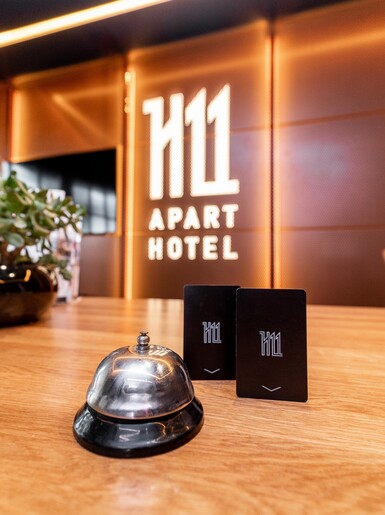 Na drewnianym blacie biurka leżą dzwonek i dwie karty hotelowe. Po lewej stoi roślina w brązowej doniczce. Na ścianie w tle znajduje się logo H11 Aparthotelu.