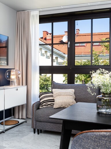 Nowoczesny salon w apartamencie. Po prawej, na czarnym stole, leżą wazon z białymi kwiatami i ozdobny talerz. Dalej, przed dużym oknem z ciemnymi ramami, znajduje się szara kanapa. Przy ścianie po lewej stoją białe szafki, a nad nimi wisi telewizor.