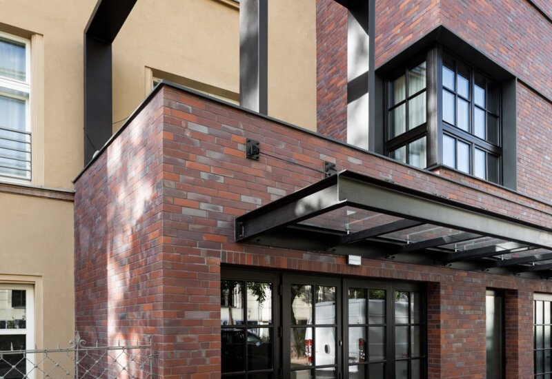 Budynek w nowoczesnym stylu ze ścianami pokrytymi cegłą. W centrum szerokie przeszklona drzwi, a nad nimi szklany daszek.