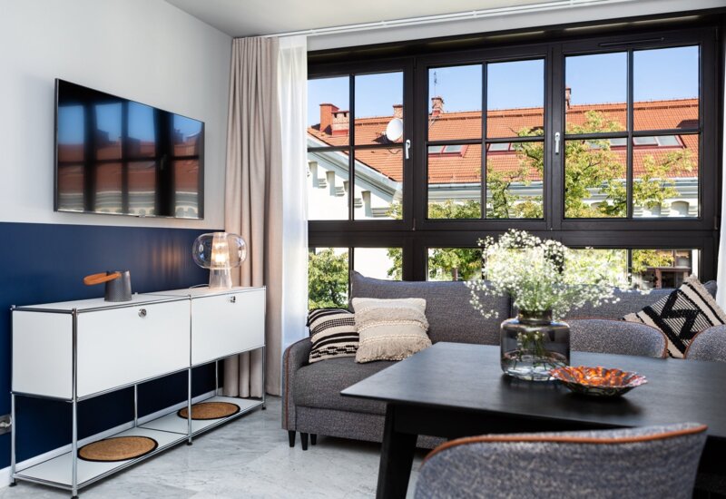 Pośrodku salonu stoi szara kanapa, przed nią stół z krzesłami. Po lewej, nad szafką wisi telewizor. Z tyłu okno na całą ścianę.