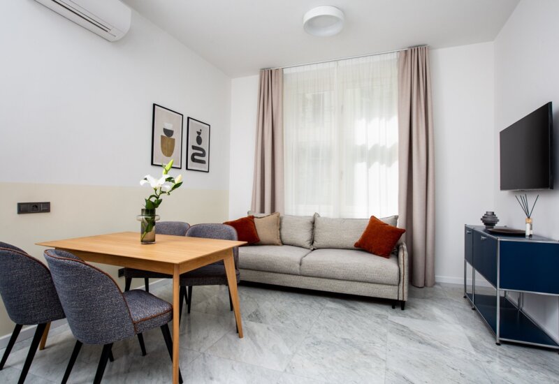 Przytulny salon w apartamencie. Po lewej stoi drewniany stół z szarymi, tapicerowanymi krzesłami. Dalej znajduje się beżowa kanapa z czerwonymi poduszkami, a za nią okno z firanką i zasłonami. Na ścianie wiszą dwie minimalistyczne grafiki.