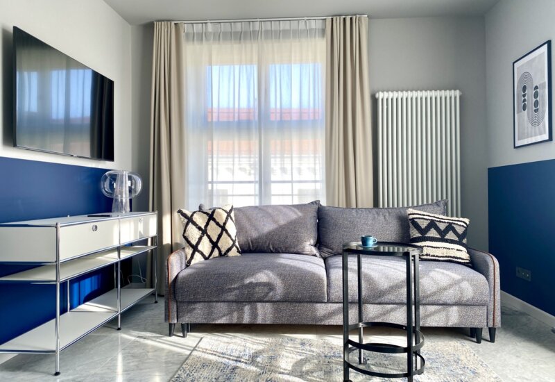 Jasny salon w apartamencie. Pośrodku szara kanapa z poduszkami. Przed nią stolik kawowy. Na ścianie po lewej telewizor, a pod nim biała szafka. z tyłu okno z firankami i zasłonami.