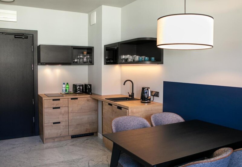 Aneks kuchenny z drewnianymi szafkami na dole i czarnymi u góry. Po prawej czarny stół z tapicerowanymi krzesłami.