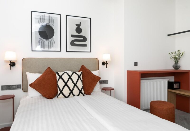 Zasłane łóżko z białą pościelą i ozdobnymi poduszkami zachowanymi w bieli, czerni i brązie. Nad zagłówkiem wiszą dwie czarno-białe grafiki. Obok szafka nocna i kinkiet.